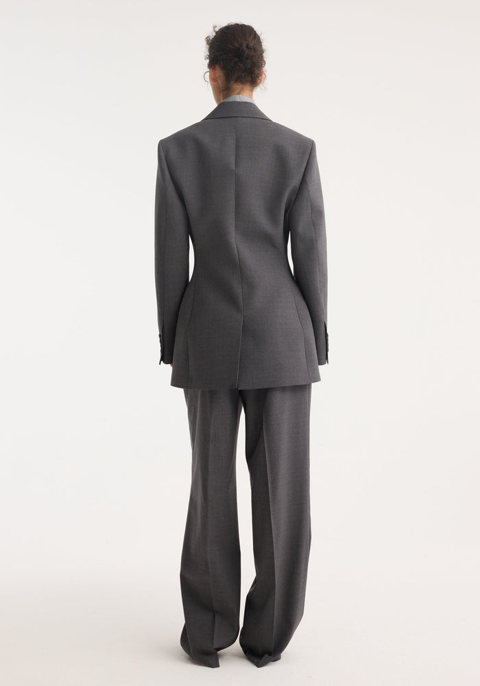 Rear view of model in Hourglass blazer, dark grey melange, showcasing a tailored fit with a sharp notch lapel, cinched waist, and wide-leg trousers.