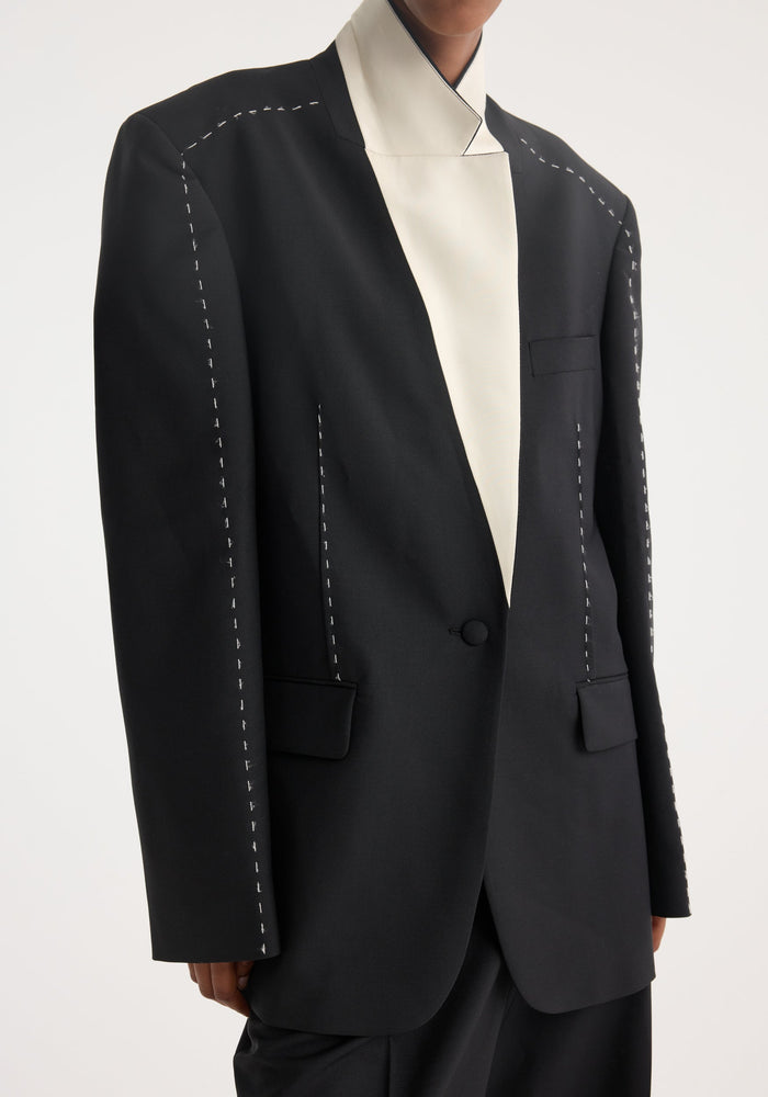 Contrast detail blazer in black, featuring oversized fit, single-button closure, white stitching accents, flap pockets, and a high-neck white top underneath, showcasing minimalist design.
