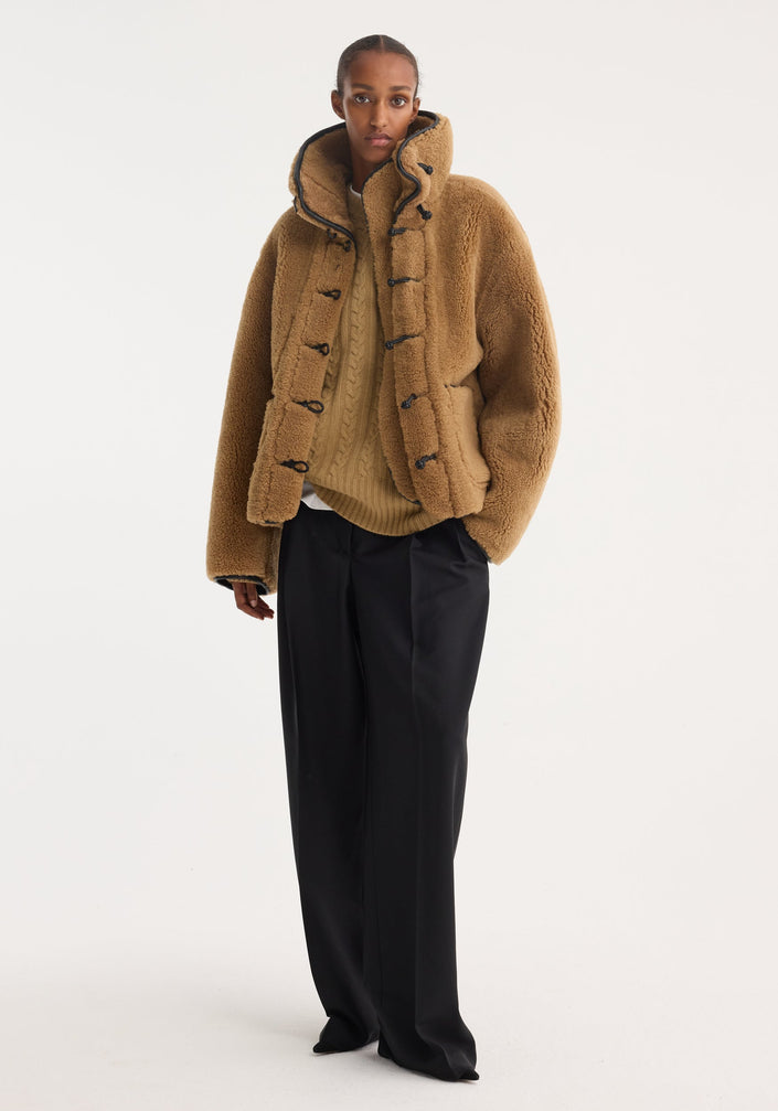 Model wearing oversized Shearling coat | cognac with high collar, toggle fastenings, patch pockets, over chunky sweater and wide-leg trousers. Modern, textured ensemble.