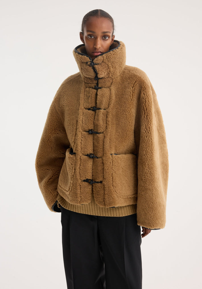Model wearing Shearling coat | cognac in plush fabric with a high collar, toggle closures, and large patch pockets, over black trousers.