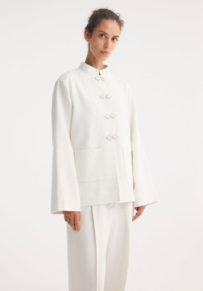 Slim model wearing the Mandarin jacket | winter white, featuring frog closures, wide sleeves, and patch pockets, paired with matching high-waisted, wide-leg trousers.
