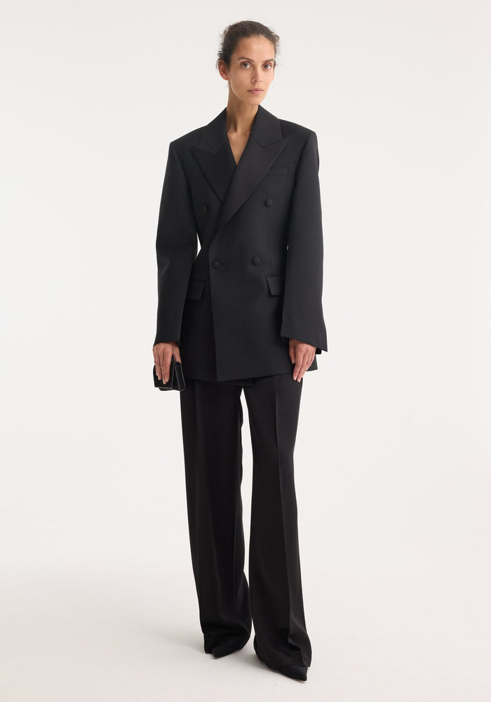 Woman wearing Hourglass tuxedo blazer with sharp notch lapels, strong shoulders, and cinched waist, paired with matching high-waisted trousers, holding a black clutch.