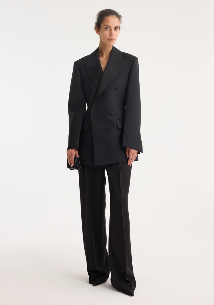 Woman wearing Hourglass tuxedo blazer with sharp notch lapels, strong shoulders, and cinched waist, paired with matching high-waisted trousers, holding a black clutch.