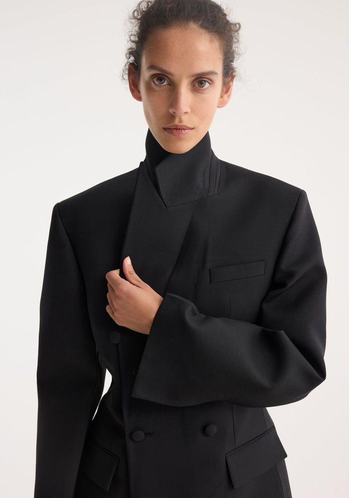 Hourglass tuxedo blazer in black with structured collar, double-breasted design, asymmetric lapel, and cinched waist, worn by model with sleek hairstyle.