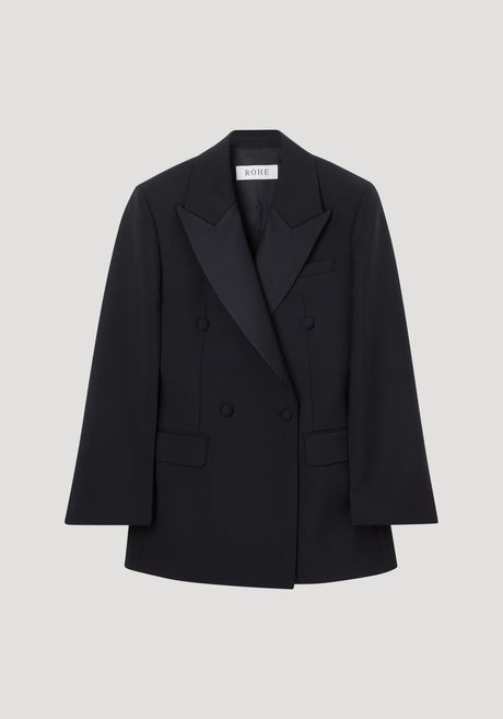 Hourglass tuxedo blazer in black with peaked satin lapels, flap pockets, and covered buttons, showcasing a sculptural silhouette with a cinched waist and strong shoulders.
