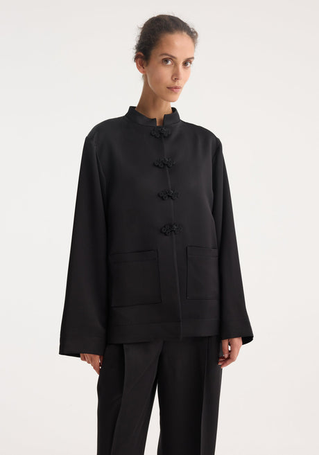 Model wearing a loose-fitting, black Mandarin satin jacket with pankou closures and patch pockets, paired with matching wide-leg trousers for a minimalist, refined look.