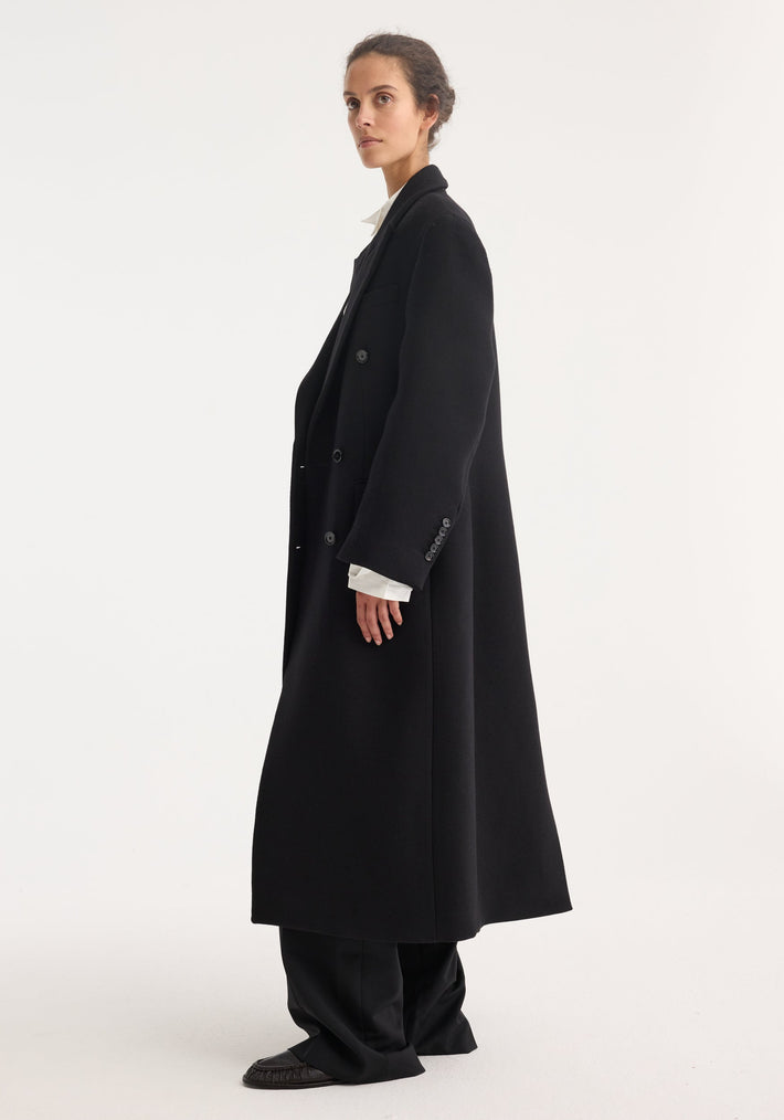Person wearing Hourglass wool coat in black, featuring a double-breasted design with strong shoulders and a feminine waist, paired with black trousers and shoes.