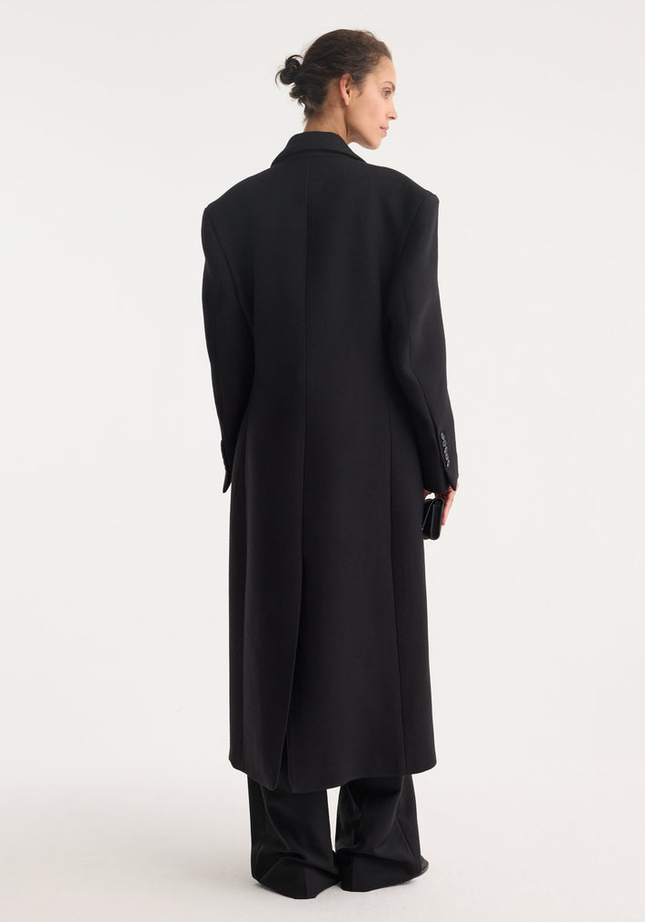 Hourglass wool coat in black, worn by model with back to camera, showcasing its long length, tailored fit, and double-breasted design paired with wide-leg trousers.