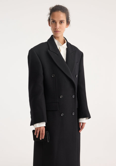 Model wears the Hourglass wool coat in black, featuring a double-breasted design with strong shoulders and a feminine waist, paired with wide-leg trousers.