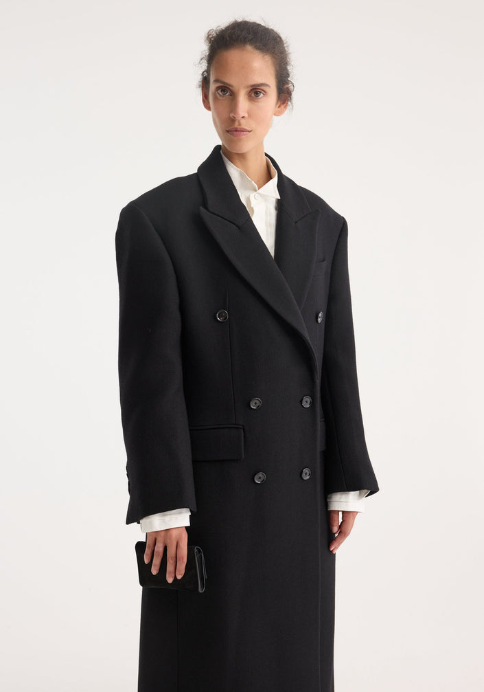 Model wears the Hourglass wool coat in black, featuring a double-breasted design with strong shoulders and a feminine waist, paired with wide-leg trousers.