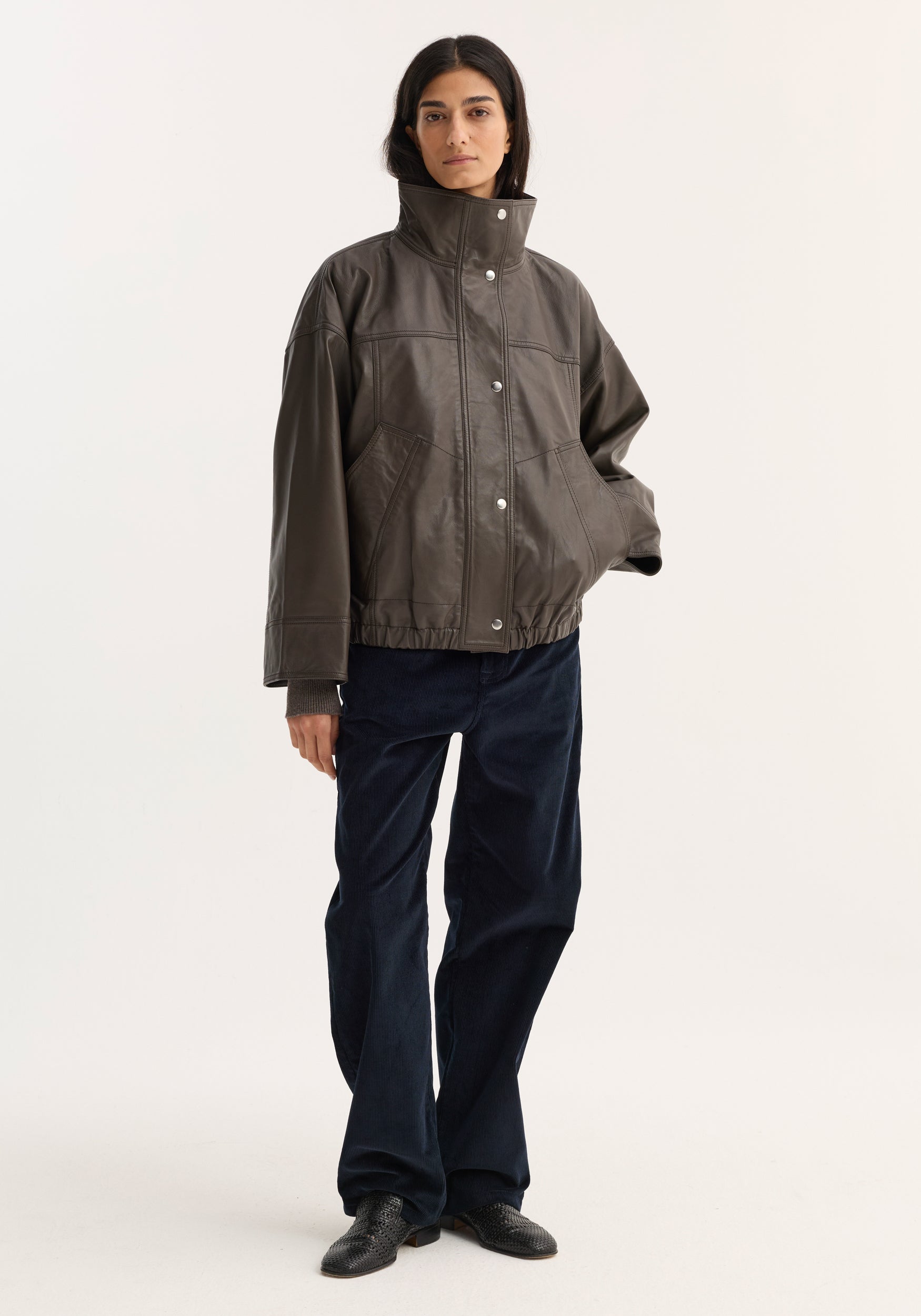 Woman wearing a dark earth-toned Reinvented leather jacket | earth with a high collar and snap button closure, paired with navy wide-leg pants and black shoes.