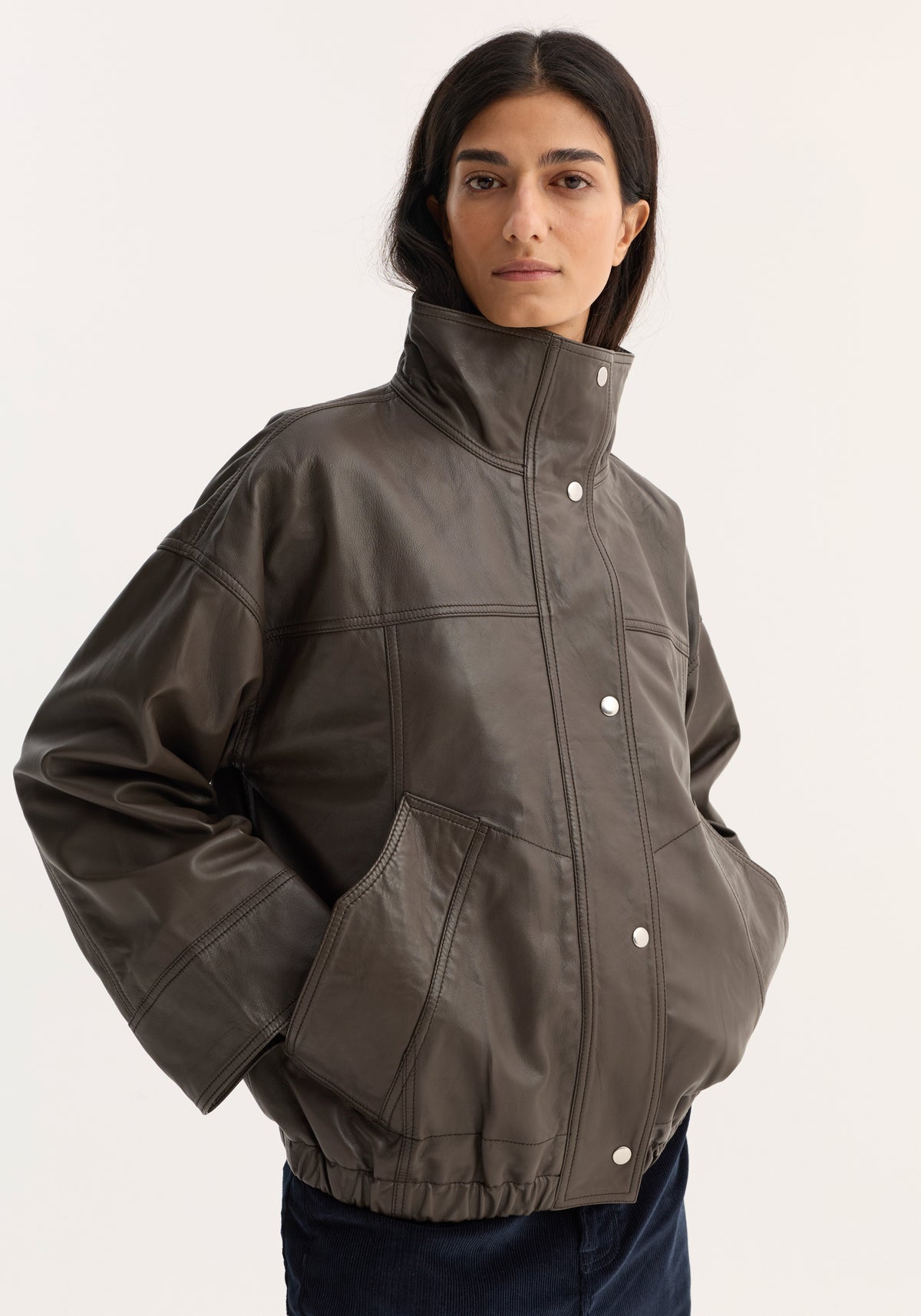 Woman wearing a dark earth-toned reinvented leather jacket | earth with a high collar, snap buttons, large front pockets, and elastic hem, posing against a plain background.