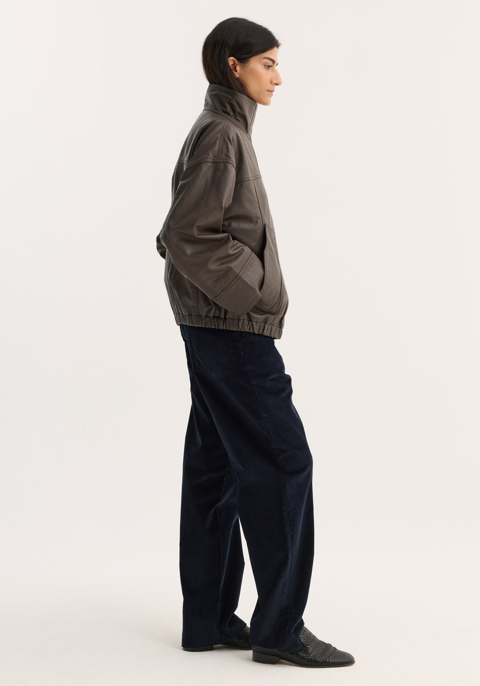 Side view of a woman wearing a high-collared, loose-fit Reinvented leather jacket | earth in a muted brown shade, paired with dark wide-leg pants and black shoes.