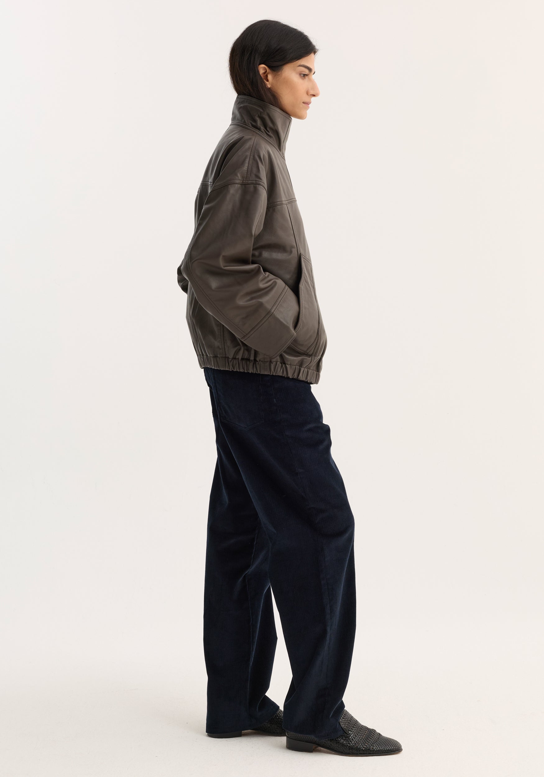 Side view of a woman wearing a high-collared, loose-fit Reinvented leather jacket | earth in a muted brown shade, paired with dark wide-leg pants and black shoes.