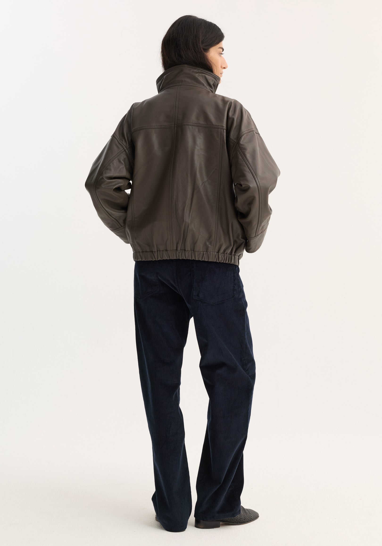 Woman seen from the back wearing a dark earth-toned Reinvented leather jacket | earth with a high collar, paired with dark blue pants and gray shoes against a plain background.