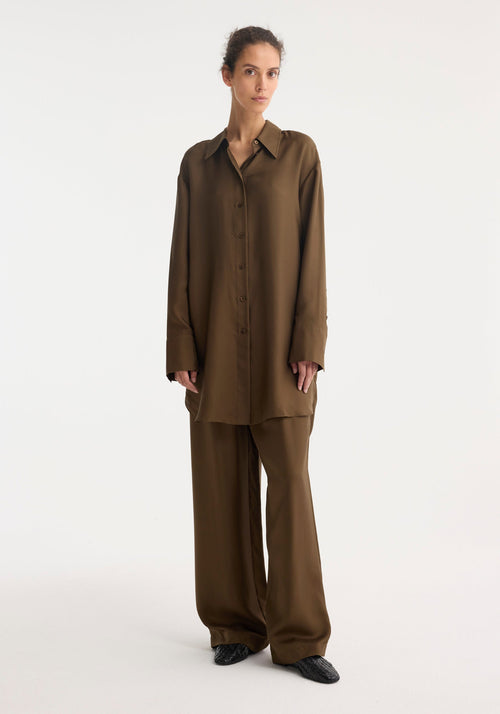 Woman wearing Signature elongated silk shirt, showcasing its oversized fit and sharp collar, paired with wide-leg trousers and slip-on shoes, embodying minimalist style.