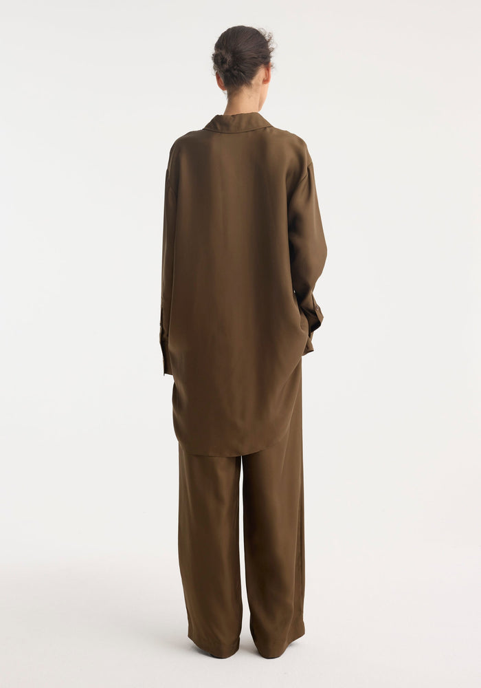 Person wearing the Signature elongated silk shirt in forest, showcasing its oversized fit and statement collar from behind, paired with matching wide-leg trousers.
