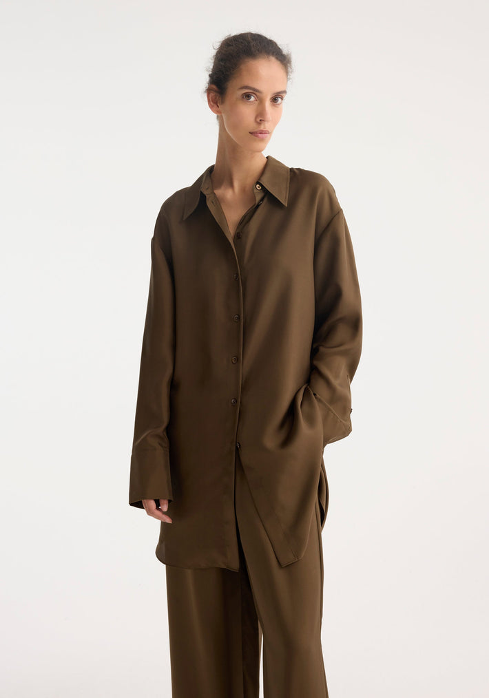 Woman wearing Signature elongated silk shirt | forest, oversized, long-sleeved, button-front, with sharp collar and cuffs, paired with matching loose-fit trousers, hand in pocket.