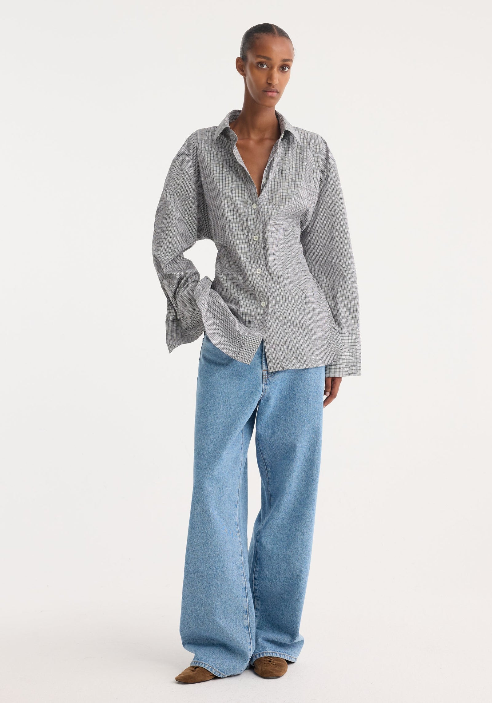 Model in a crushed checked shaped shirt with mini grey check, untucked, oversized sleeves, paired with wide-leg jeans and pointed-toe shoes, showcasing casual elegance.