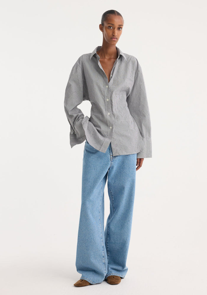 Model in a crushed checked shaped shirt with mini grey check, untucked, oversized sleeves, paired with wide-leg jeans and pointed-toe shoes, showcasing casual elegance.