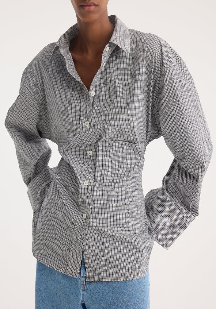 Model in a crushed checked shaped shirt with mini grey check, untucked, oversized sleeves, paired with wide-leg jeans and pointed-toe shoes, showcasing casual elegance.