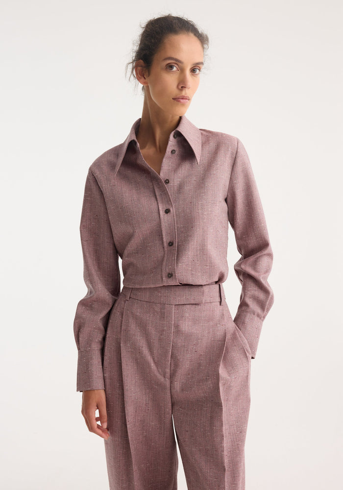 Model in Fitted Tweed Shirt | Mauve Melange, a long-sleeve shirt with pointed collar and dark buttons, paired with high-waist pleated trousers, hand in pocket.