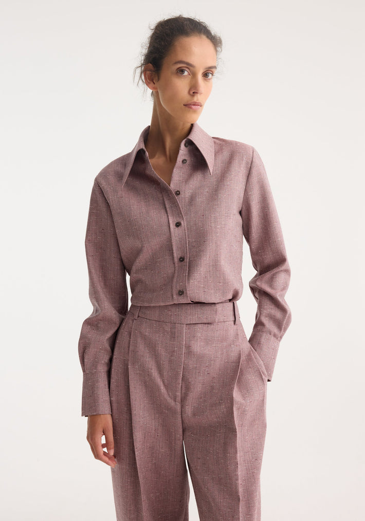 Model in Fitted Tweed Shirt | Mauve Melange, a long-sleeve shirt with pointed collar and dark buttons, paired with high-waist pleated trousers, hand in pocket.