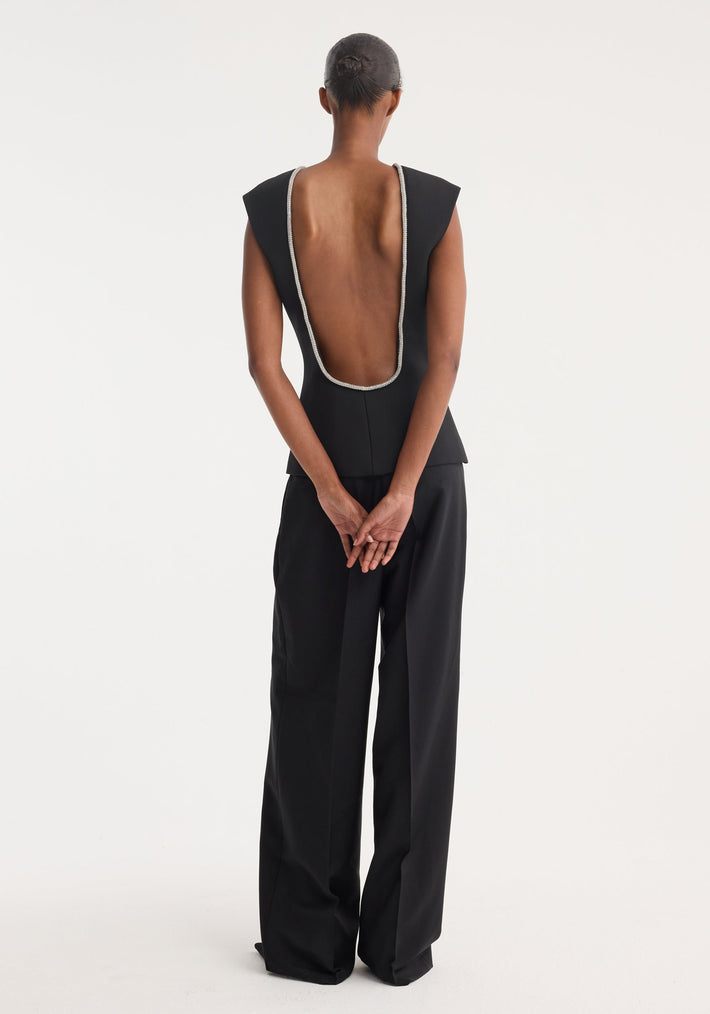 Woman in a black Open back appliqué top with deep scoop back and crystal trim, styled with wide-leg trousers, showcasing a refined silhouette.