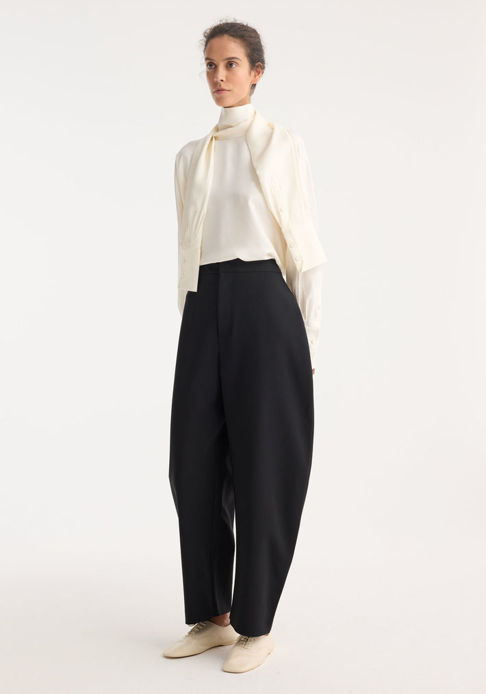 Young woman wearing sculptural trousers with a high waist and balloon legs, paired with an ivory blouse and knit, showcasing a minimalist, monochromatic look.