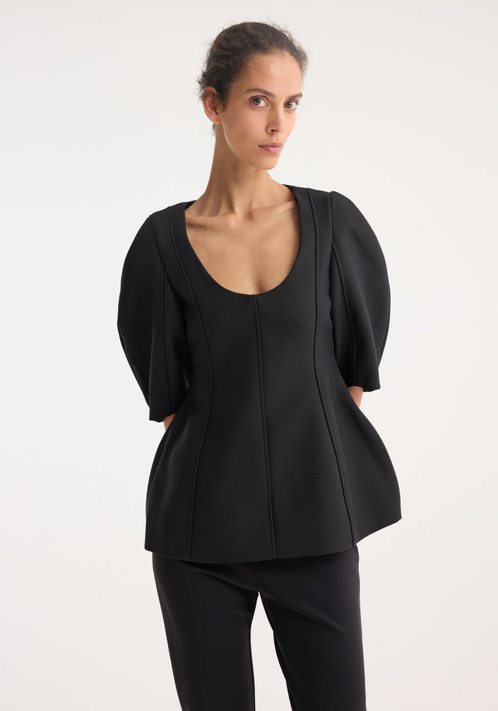 Model wearing Sculptural top | black with a deep scoop neckline, elbow-length balloon sleeves, and a peplum flare, paired with slim-fit trousers and open-toe sandals.