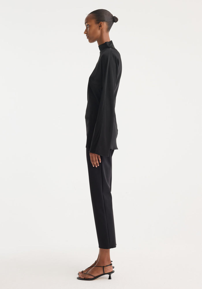 Woman in a side-profile wearing a Satin waisted top with a mock neck and flared cuffs, paired with slim trousers and strappy sandals.