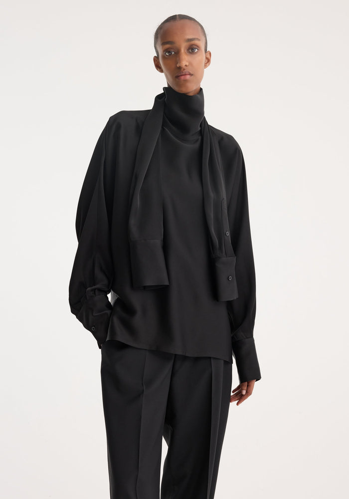 Model wearing Foulard satin top with draped sleeves, extended turtleneck as a scarf, paired with tailored trousers, showcasing a minimalist, modern silhouette.