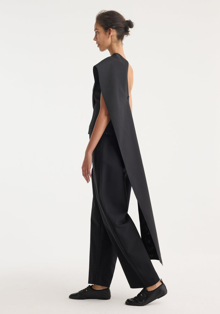 Model in profile wearing a black one-shoulder cape top with a dramatic back panel, paired with high-waisted trousers and lace-up flats.