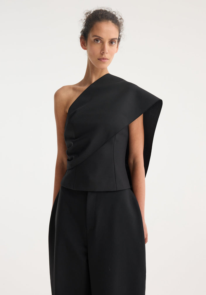 Woman wearing a One-shoulder cape top in black with an asymmetric sash drape, paired with wide-leg trousers, showcasing minimalist, contemporary fashion.