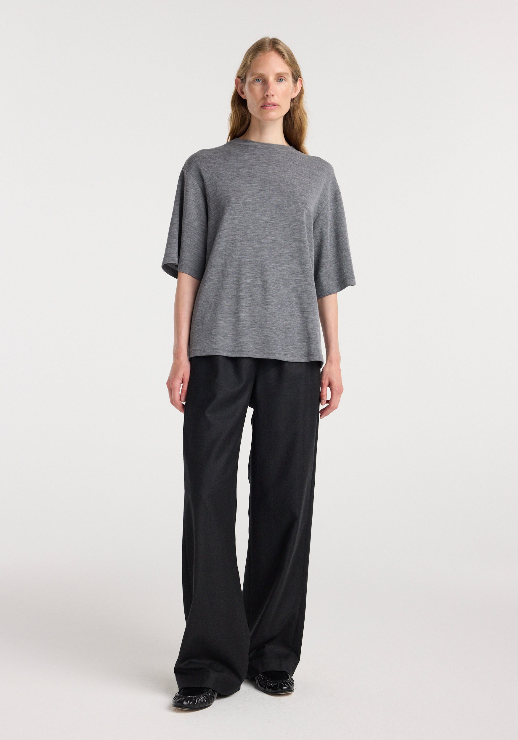 Woman wearing an oversized soft merino t-shirt | mid grey melange with wide-leg black trousers and black shoes, standing against a plain light background.