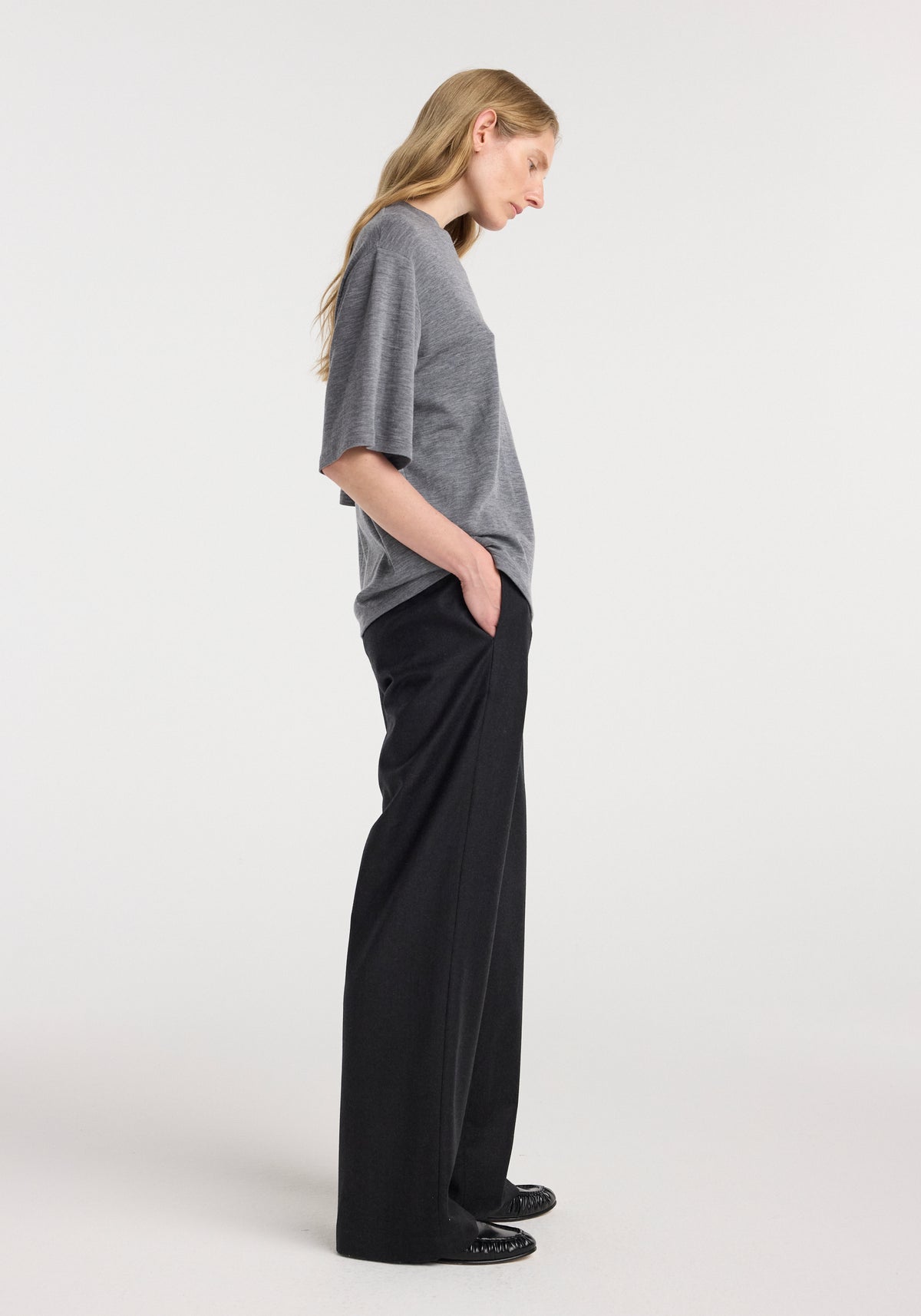 Woman wearing an oversized soft merino t-shirt | mid grey melange with elbow-length sleeves paired with wide-leg black trousers, standing in profile with hands in pockets.