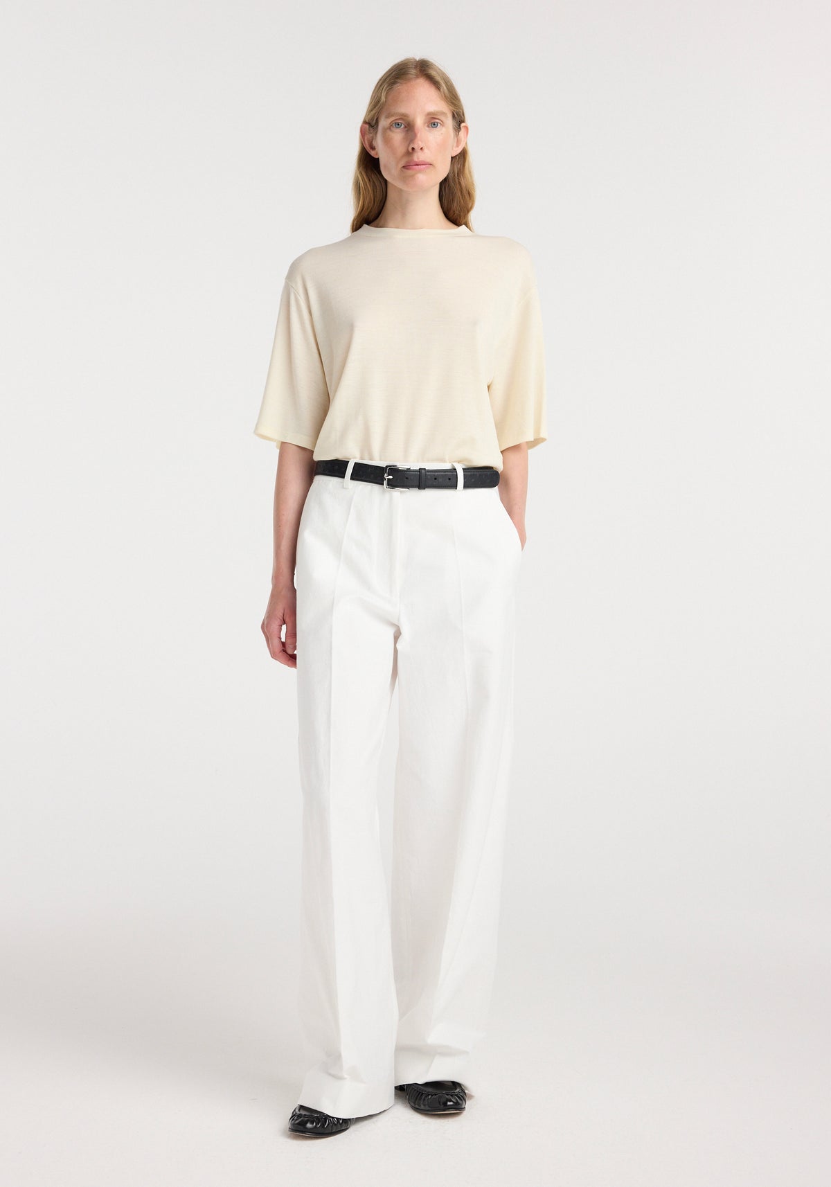 Woman wearing an oversized merino t-shirt | off white with short sleeves, paired with high-waisted white trousers and black shoes, standing against a plain background.
