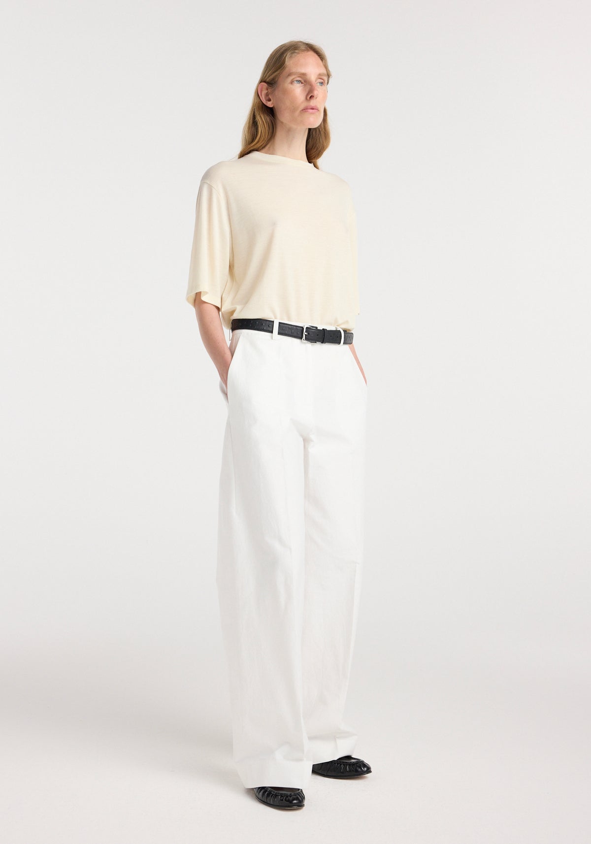 Woman standing wearing an Oversized merino t-shirt | off white paired with high-waisted white wide-leg pants, black belt, and black shoes against a plain background.