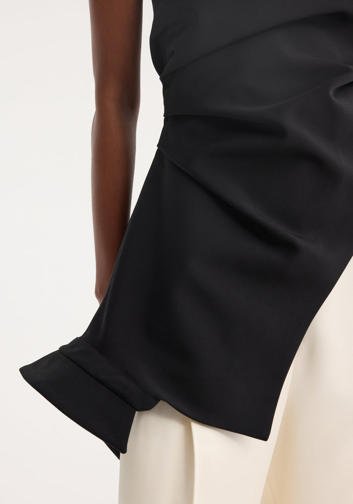 Close-up of a Reimagined one shoulder top in black, showcasing its asymmetrical drape, deconstructed detail, and upside-down collar over cream trousers.