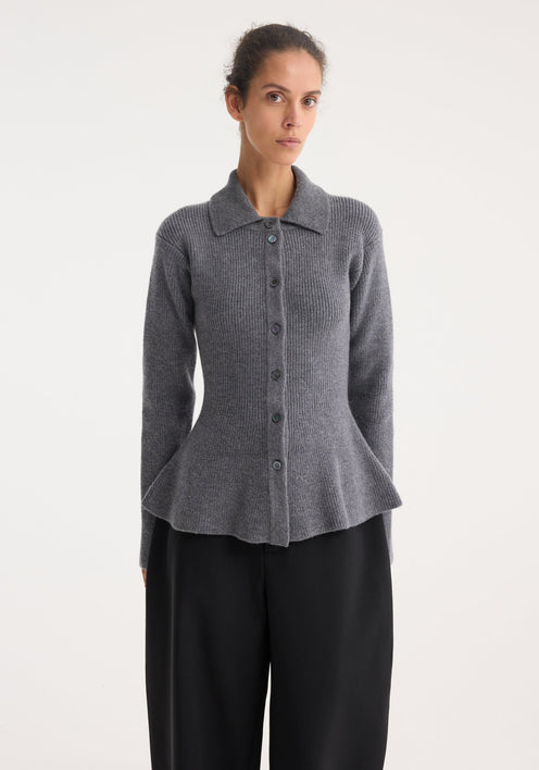 Model wearing a waisted rib cardigan in dark grey melange, featuring a peplum flare and polo collar, paired with wide-leg black trousers and lace-up shoes.