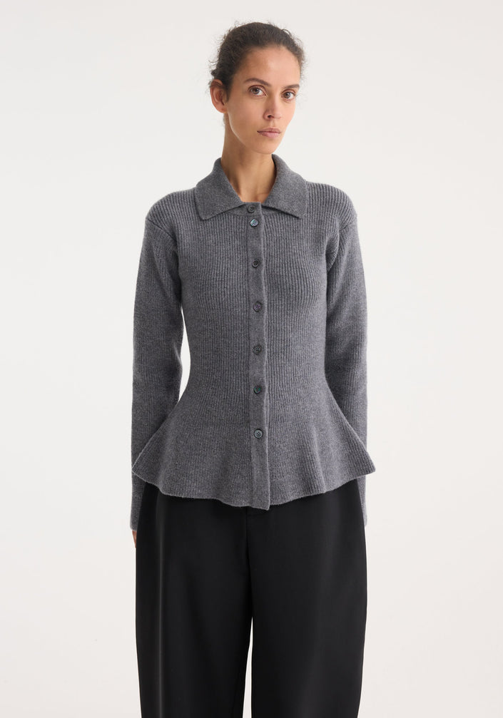 Model wearing a waisted rib cardigan in dark grey melange, featuring a peplum flare and polo collar, paired with wide-leg black trousers and lace-up shoes.