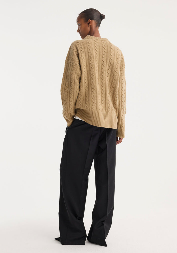 Person wearing oversized Cable knit jumper in camel, featuring dropped shoulders and a round neckline, paired with wide-leg trousers, viewed from behind.