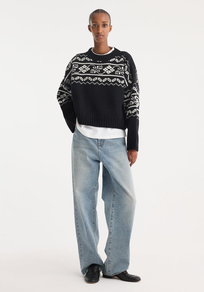 Model wearing a Fair-isle knitted jumper in black, featuring a boxy, cropped fit. Styled with light-wash wide-leg jeans and black pointed-toe shoes.