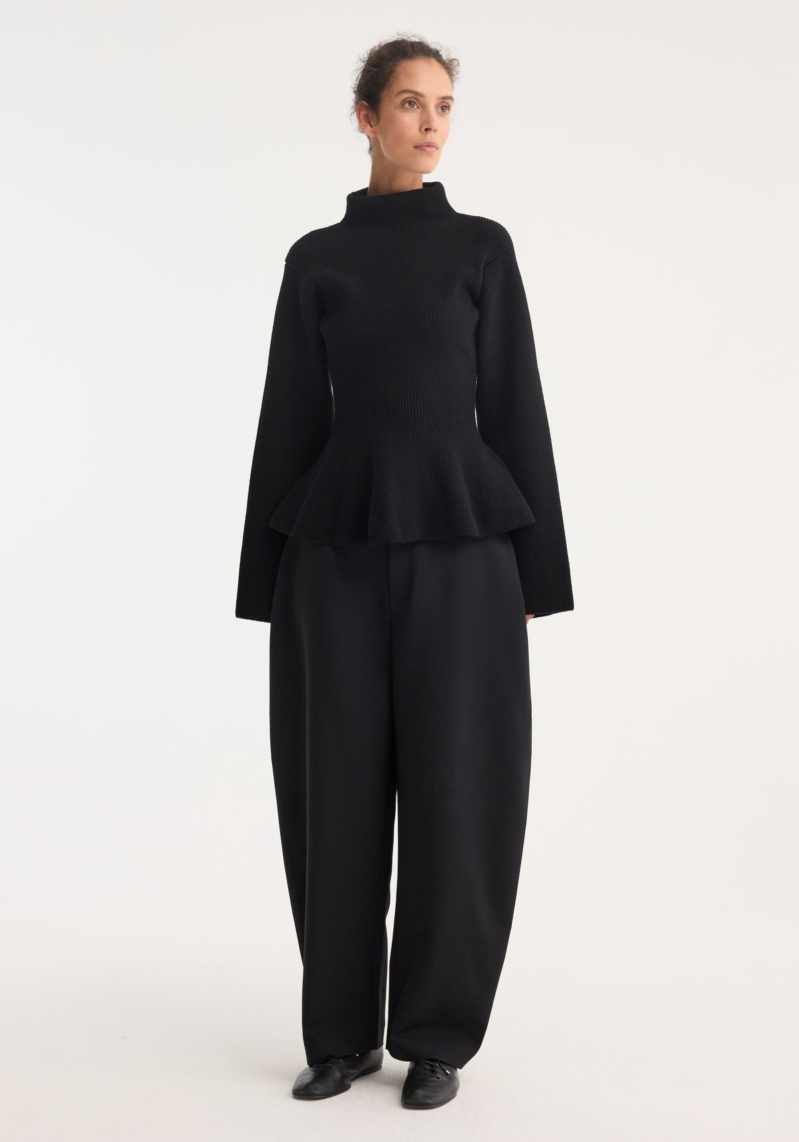 Woman wearing Sculptural knitted top with high collar, elongated sleeves, and peplum hem, paired with wide-leg trousers and lace-up shoes, gazing sideways.