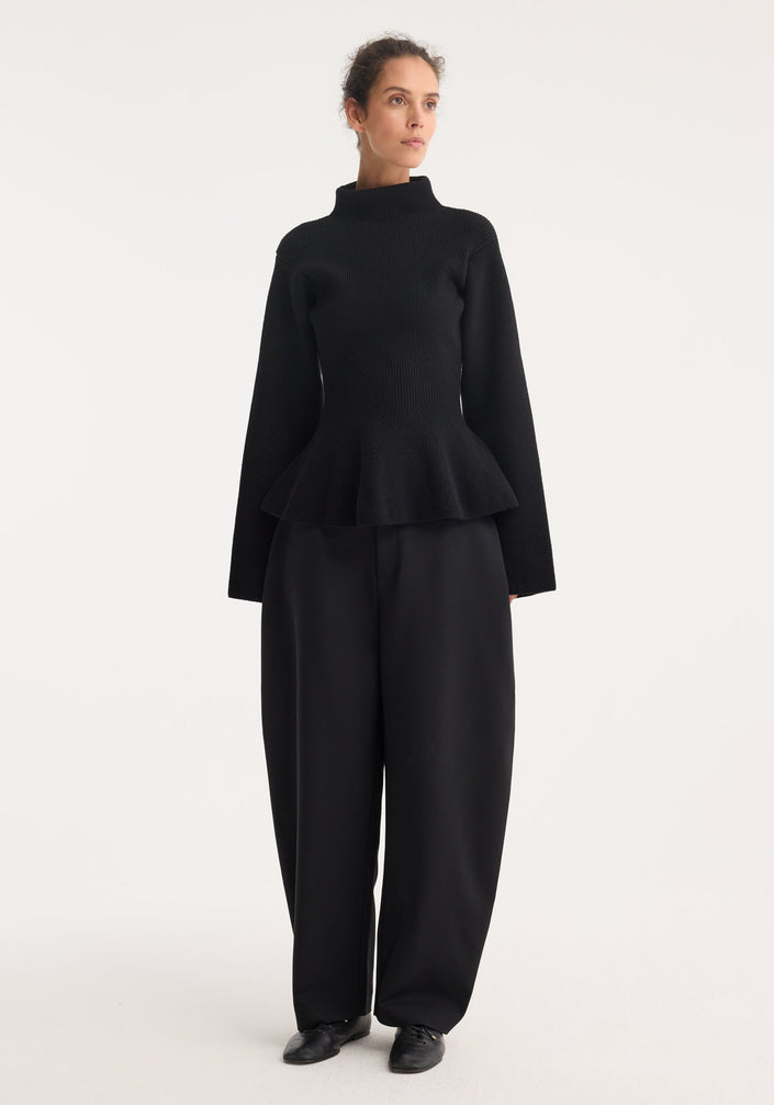 Woman wearing Sculptural knitted top with high collar, elongated sleeves, and peplum hem, paired with wide-leg trousers and lace-up shoes, gazing sideways.