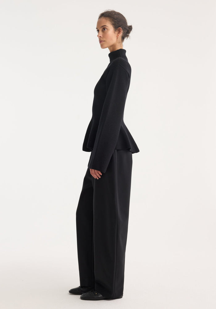 Model in side-profile wearing a Sculptural knitted top with a high collar, elongated sleeves, and peplum flare, paired with wide-leg trousers.