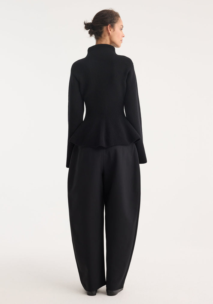 Woman wearing Sculptural knitted top with high collar, elongated sleeves, and peplum flare, paired with wide-leg trousers, viewed from behind.