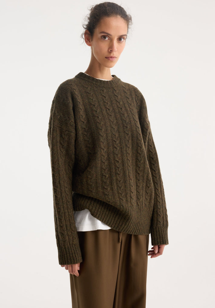 Woman wearing Cable knit jumper | forest melange, oversized wool pullover with cable texture, crew neckline, and dropped shoulders, layered over a white T-shirt.