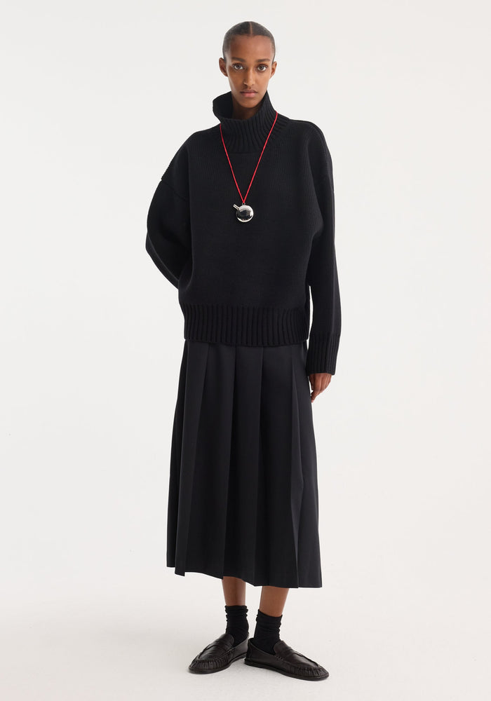 Model in sculptural compact turtleneck, black ribbed with dropped shoulders, paired with pleated skirt and loafers, accessorized by metallic pendant on red cord.