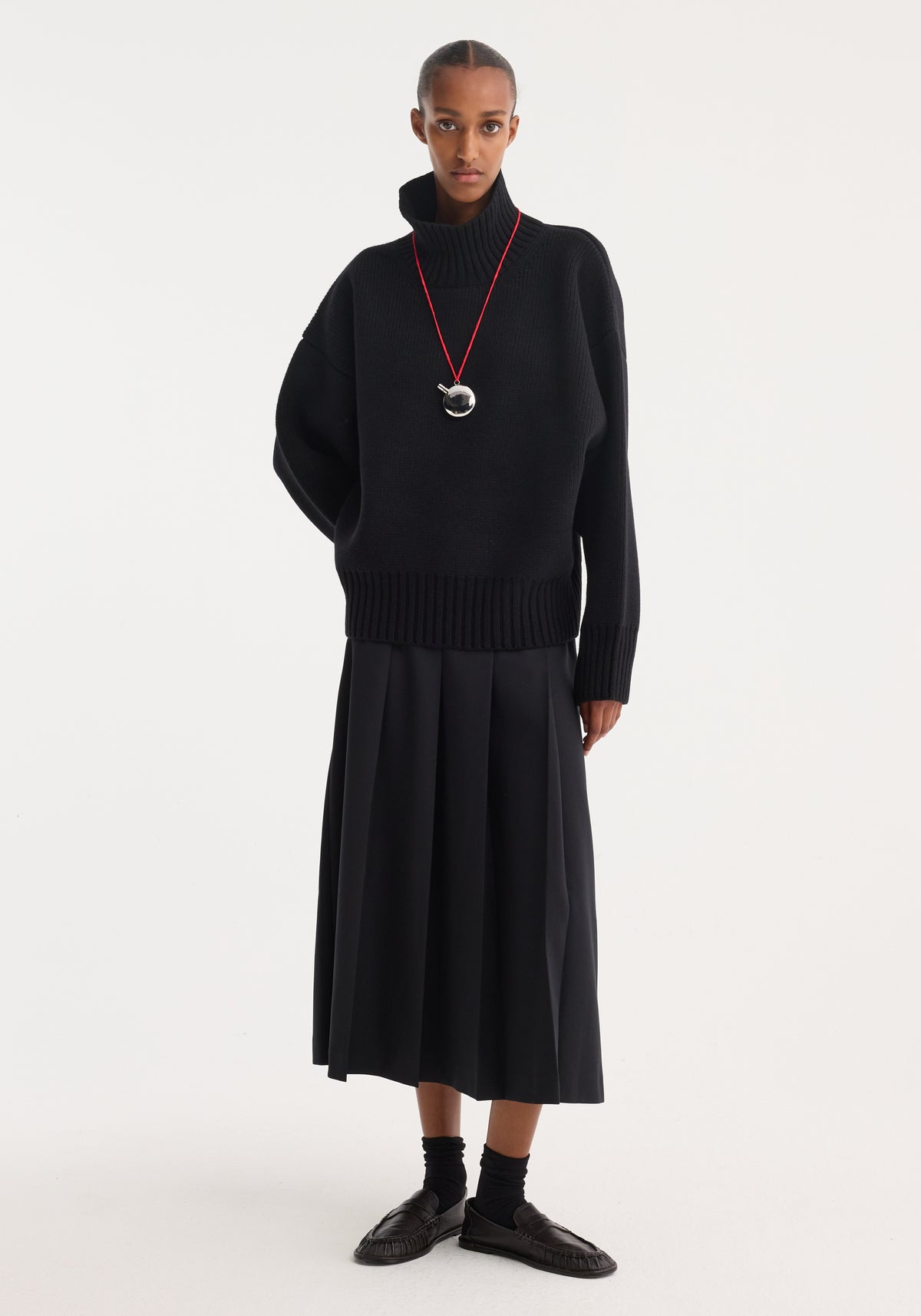 Model wearing a black sculptural compact turtleneck | black with long sleeves, paired with a pleated black midi skirt, black socks, and black shoes, accessorized with a necklace featuring a silver pendant on a red cord.